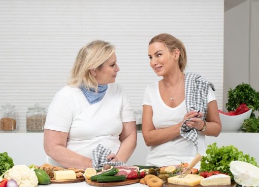 Two women in a kitchen preparing fresh vegetables and cheese with olive oil and spices.